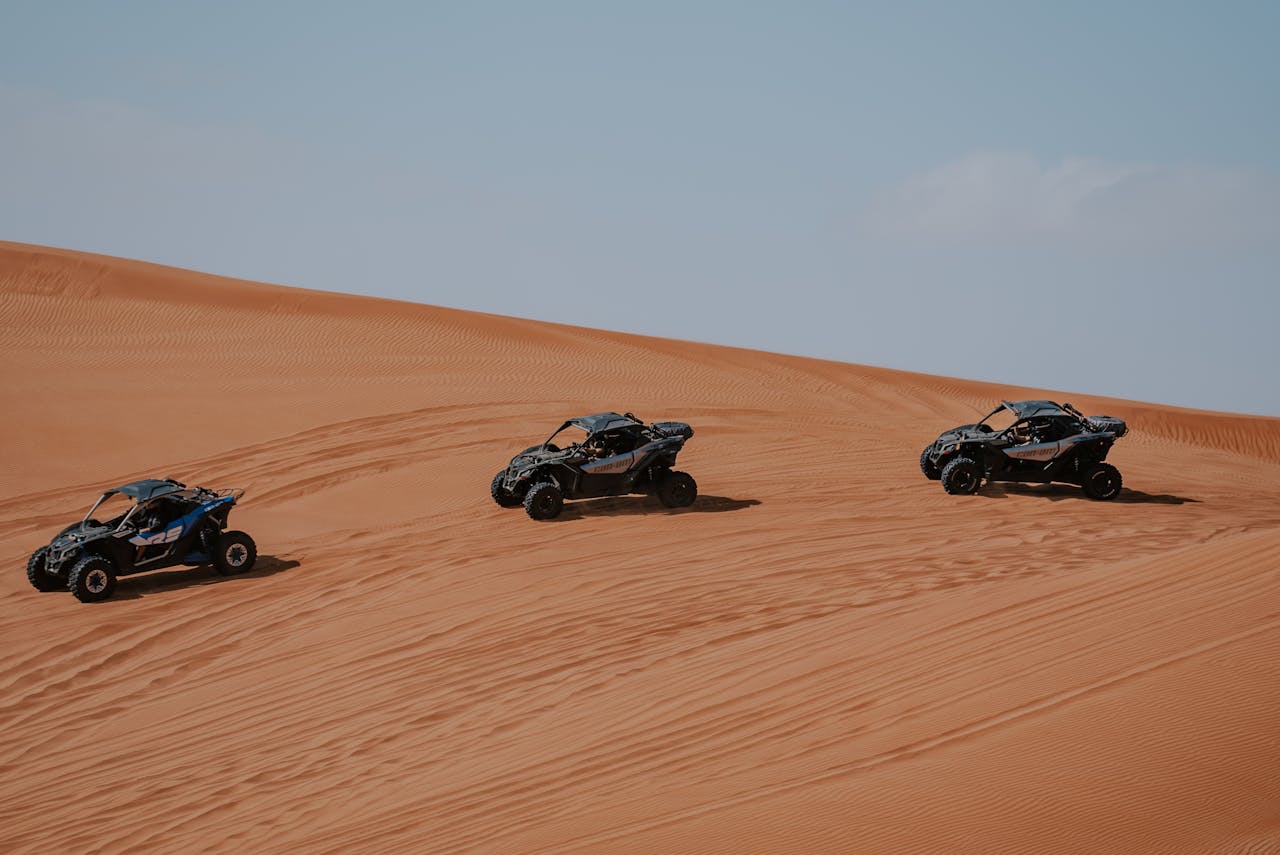 Three all-terrain vehicles racing across expansive desert sand dunes under a clear sky.
