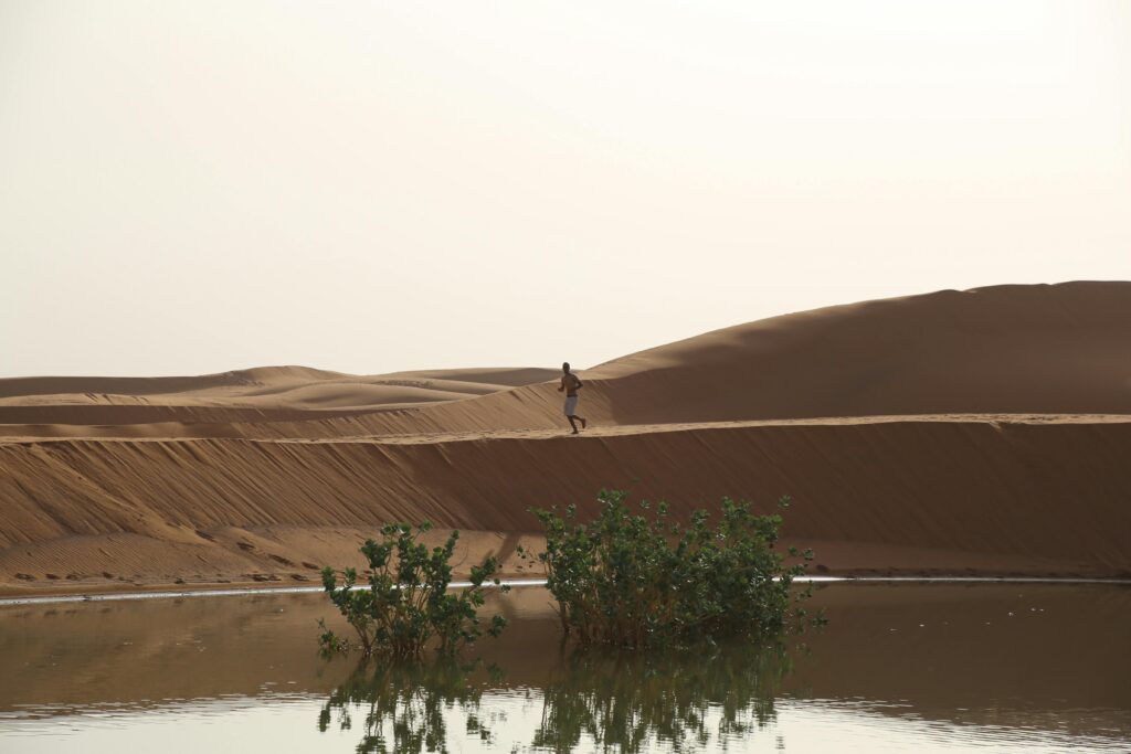 A lone man walks across sandy dunes near a reflective pond in Merzouga, Morocco, showcasing desert beauty.